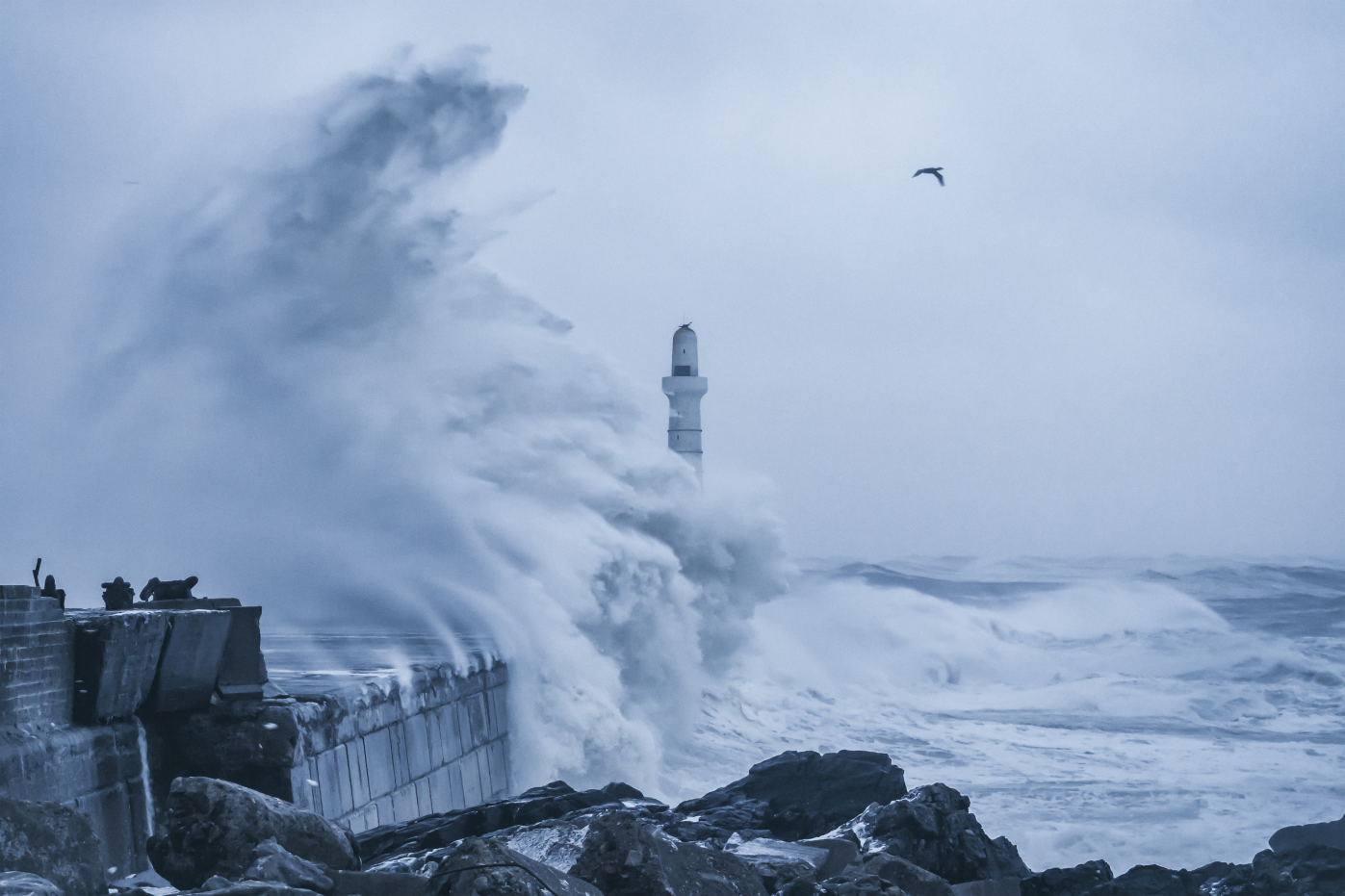 A powerful wave crashes over a pier and lighthouse, displaying the force of nature's might and the vulnerability of man-made structures.